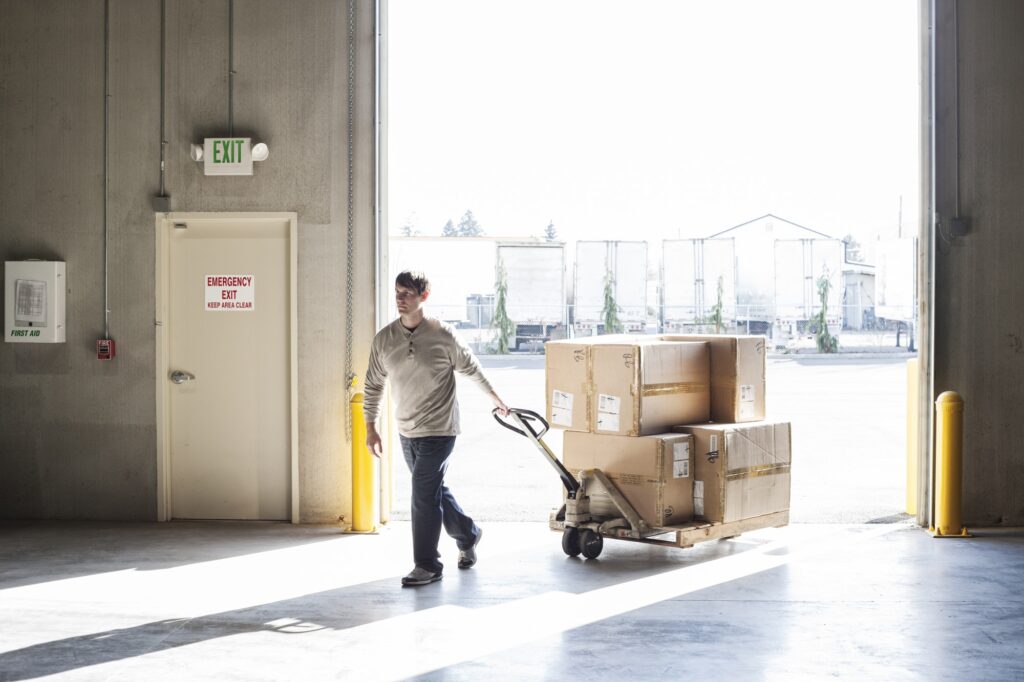 A male warehouse worker moving boxes of products using a manual pallet jack.